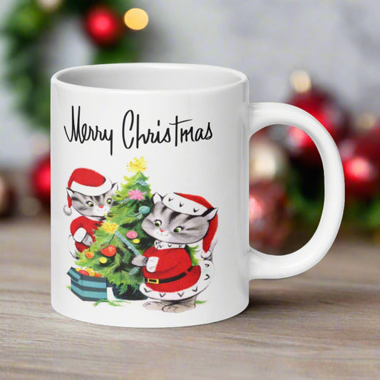 This glossy ceramic coffee mug features a vintage illustration of two cats in santa outfits decorating their Christmas tree. It says Merry Christmas above the artwork in a Midcentury style lettering.