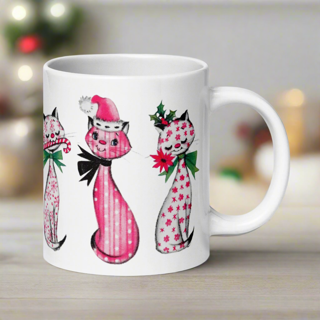 This ceramic glossy coffee mug features a midcentury retro Christmas illustration of three cats. They are adorned with bows around their necks. One has a pink santa hat on her head, one has a candy cane in her mouth and the other is holding a flower.